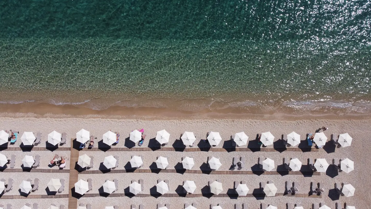 Aerial view of a Mediterranean beach club: rows of white parasols on pale sand beside clear turquoise water